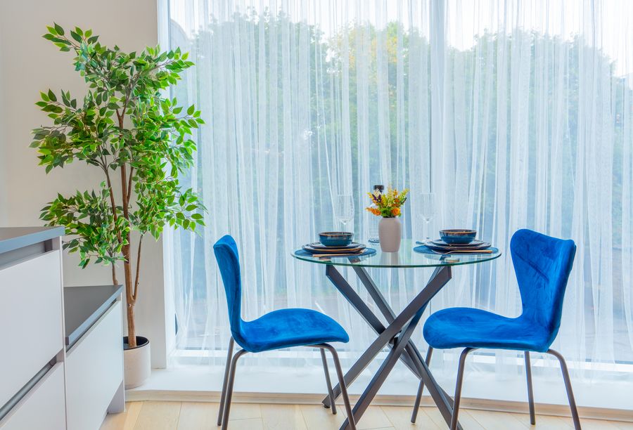 Round glass dining table with two blue chairs by the window